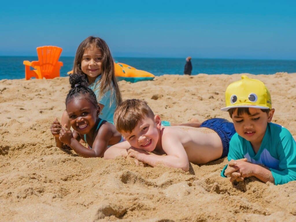 An authentic, real photography shot of a group of diverse children smiling and lying in the sand at the beach on a sunny day.