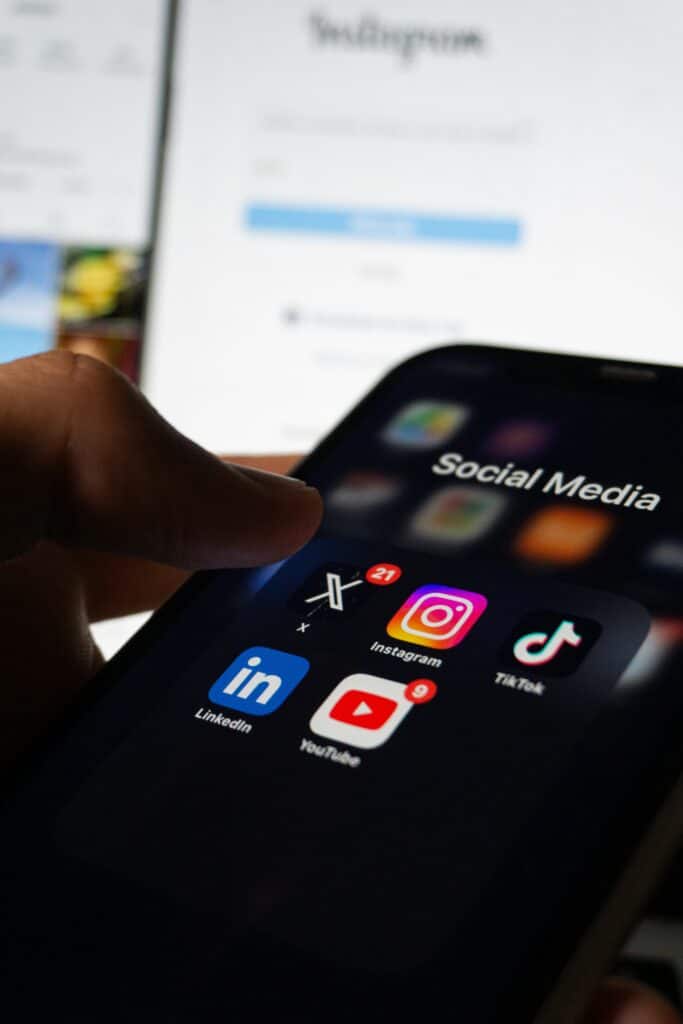 A close-up of a person's thumb navigating social media app icons—including Instagram, X, TikTok, LinkedIn, and YouTube—on a smartphone screen.