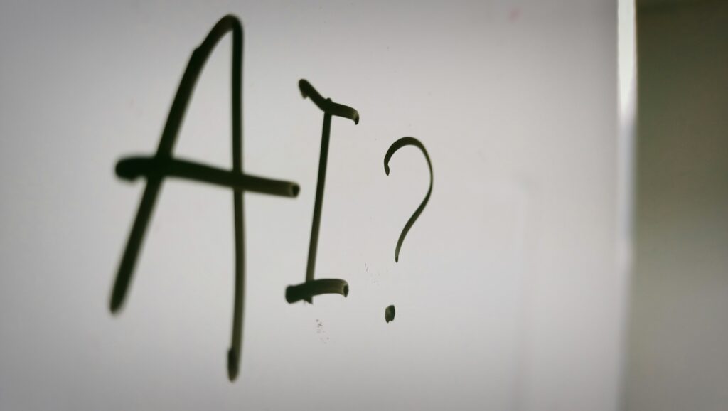 The letters AI followed by a question mark handwritten in black marker on a white gloss surface, symbolizing uncertainty in artificial intelligence strategy.