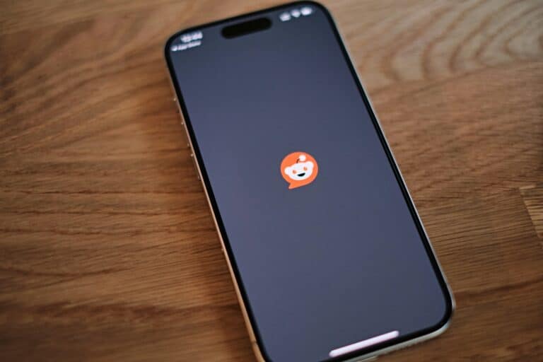 An iPhone lying on a wooden table screen-side up, displaying the orange and white Reddit mobile app logo in the center of a dark screen.