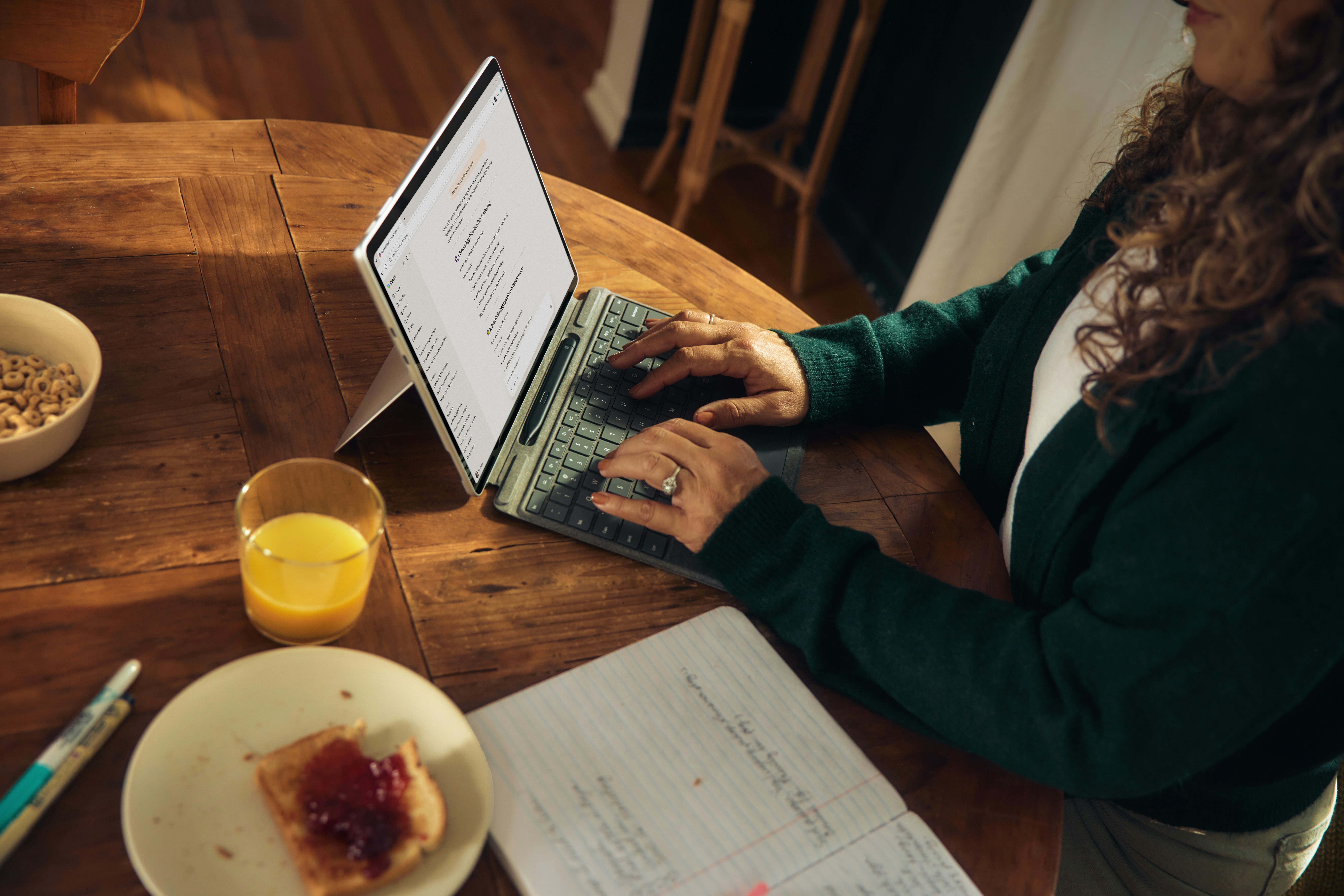 An over-the-shoulder view of a woman typing on a 2-in-1 laptop tablet at a table with a notebook nearby, highlighting content structure for SEO and LLMO.