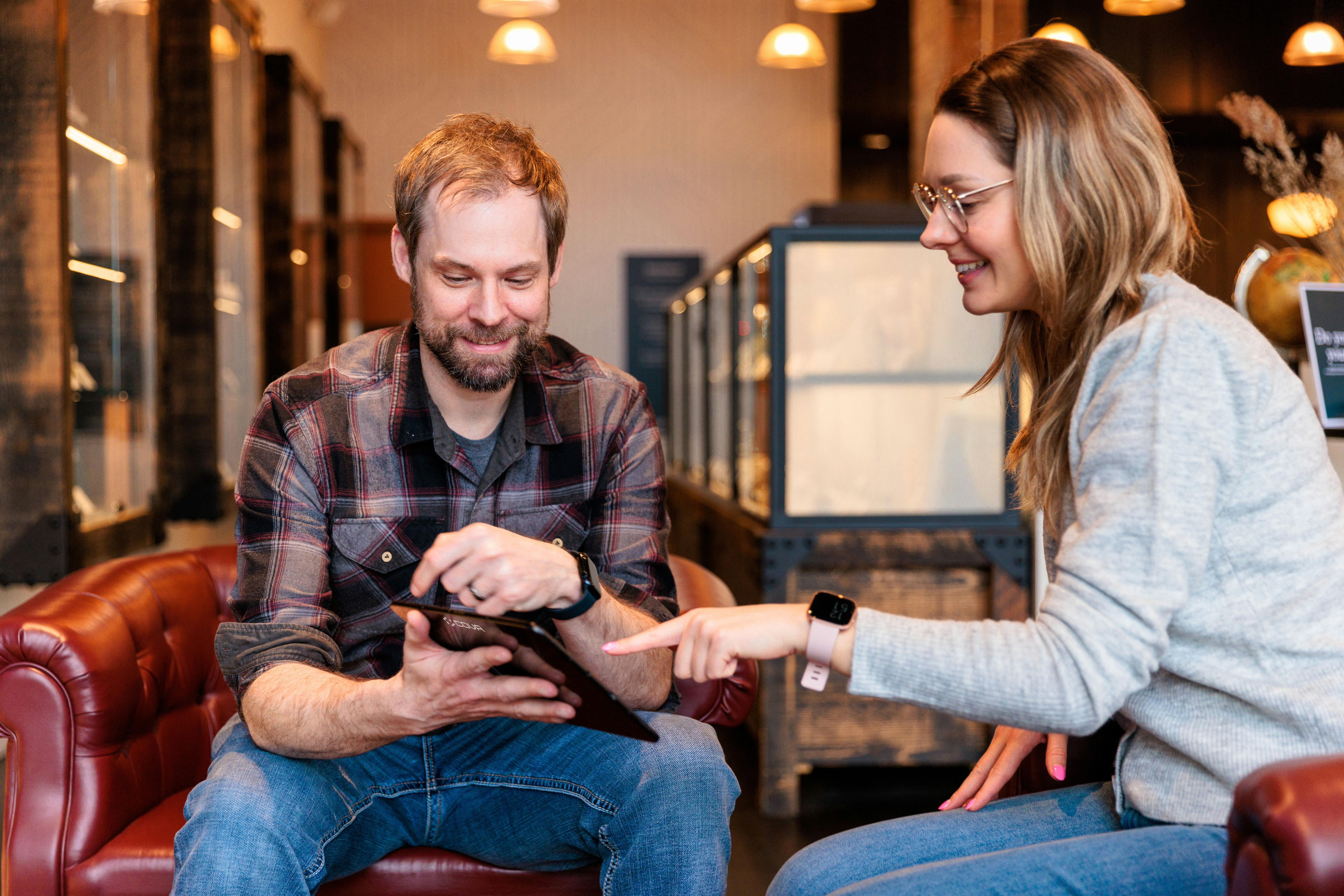 Smiling man and woman sitting in leather armchairs pointing at a tablet screen together.