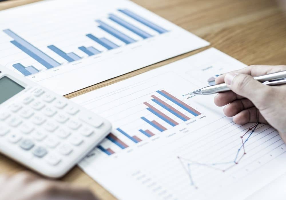 Person pointing a pen at financial performance bar graphs and chart documents on a wooden desk with a white calculator.