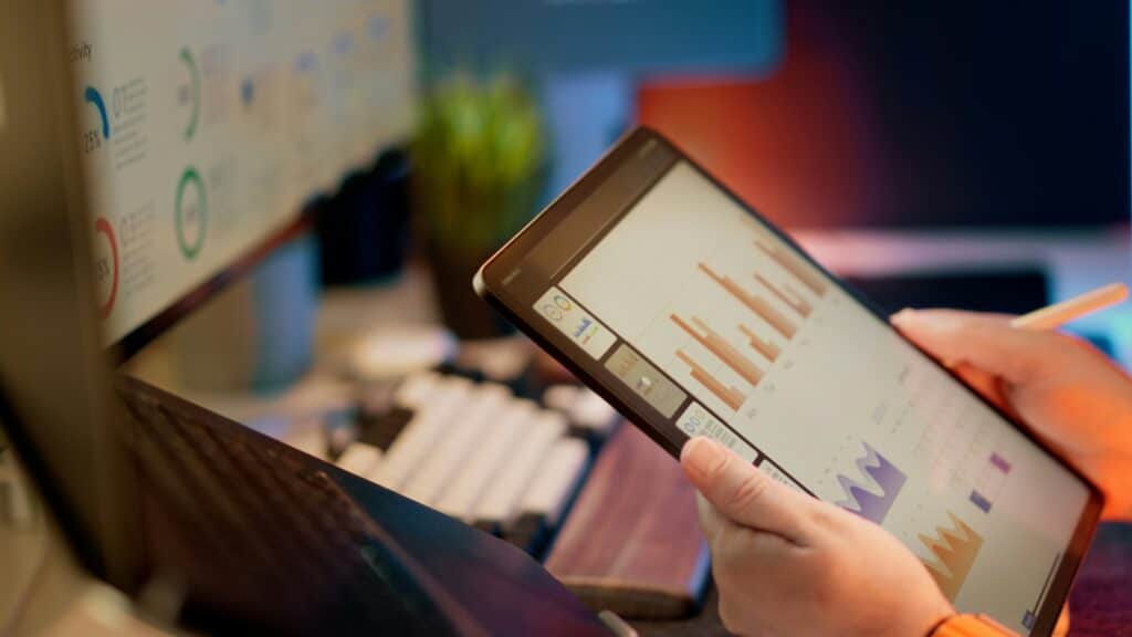 Close-up of hands holding a tablet displaying color bar charts and analytics dashboards next to a desktop screen.