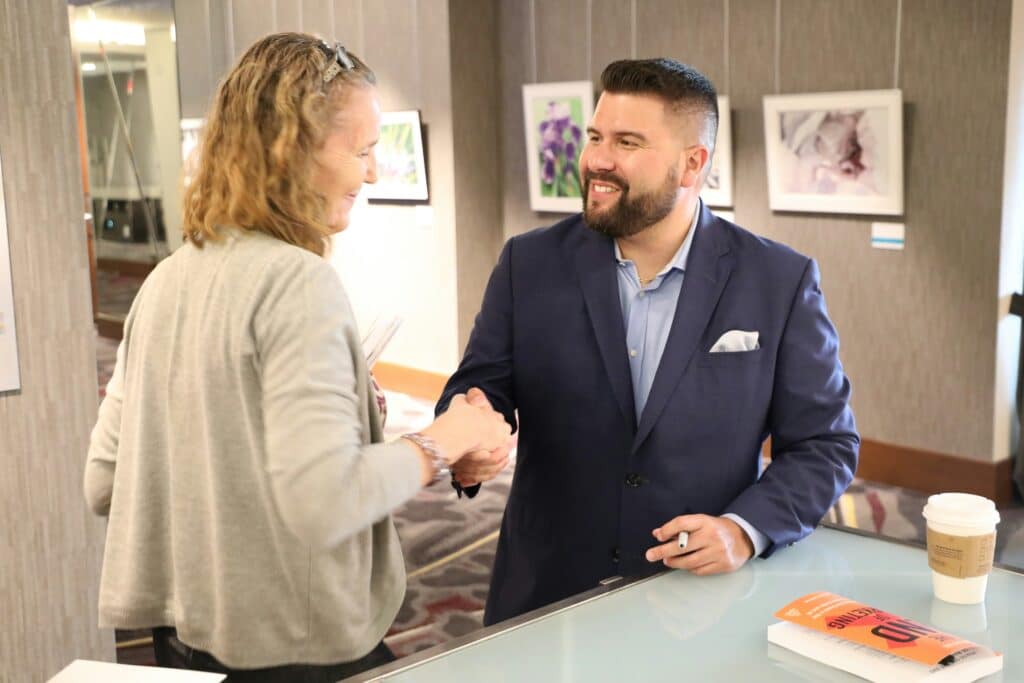 A smiling businessman in a navy suit jacket shaking hands with a client across a counter.